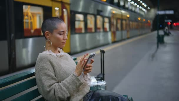 移動式携帯電話の出発スケジュールを見ている観光ステーションベンチを閉じます 電車を待っている携帯電話のアプリケーションを閲覧するケアフリーの短い髪の女性 リラックスしたトレンディな少女の航海の読書メッセージ — ストック動画