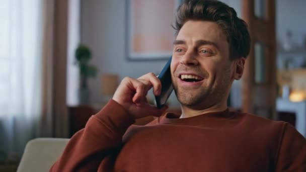 ハッピーマンは クローズアップして座っているアパートのソファーを楽しんでいます 週末にスマートフォンで話す魅力的な幸せな男の肖像画 笑顔のハンサムなヒップスター 屋内の携帯電話の会話でニュースを共有 — ストック動画