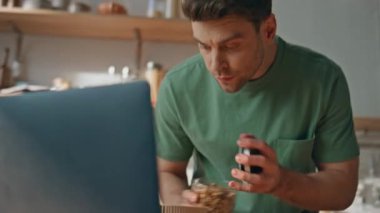 Modern apartman dairesinde tabağa granola döken esmer kadın. Dizüstü bilgisayar seyreden traşsız bir adam hafif mutfakta kahvaltı hazırlıyor. Sakin adam bilgisayar ekranında yalnız zaman geçiriyor.