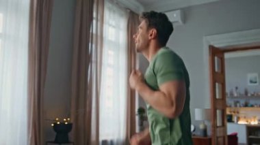 Enerjik sporcu evde egzersiz yapıyor. Yakın çekim. Hevesli genç adam modern oturma odasında kardiyo egzersizleri yaparak zıplıyor. Yakışıklı kaslı adam apartmanın içinde sabah sporu yapıyor..