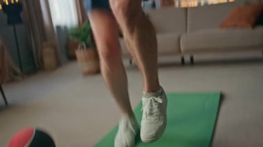 Sporcu ayakları, yakın planda spor minderinde aktif olarak zıplıyor. Oturma odasında egzersiz yapan genç ve güçlü bir adam sağlıklı bir vücut geliştiriyor. Motivasyonlu yakışıklı sporcu dairedeki günlük antrenmandan önce ısınıyor.