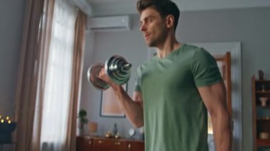 Apartman dairesinde kaslı atletlerin kol kaslarını çalıştırması. Yakın çekim. Ciddi motivasyona sahip adam evde güçlü eller için dambılları kaldırıyor. Yakışıklı sporcu içeride ekipmanla spor yapıyor..
