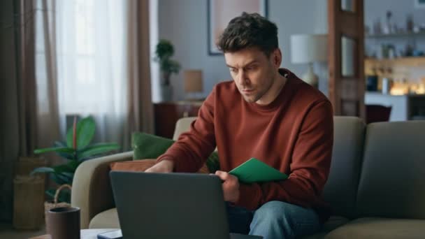 坐在舒适的沙发上 坐在笔记本电脑前观看在线课程的家伙 一个严肃的男人在家里喝着咖啡 以俊俏的学生为主要对象 在现代公寓里拿着电脑显示屏 — 图库视频影像