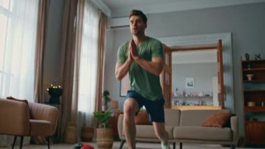 Atletizm antrenmanı evde. Modern bir apartman dairesinde paspas eğitimi veren vücuda odaklanmış güçlü adam hamlelerini yapıyor. Formda tişörtlü sporcu bacak kaslarını güçlendiriyor..