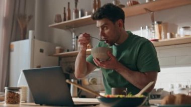 Kahvaltıda laptopunu okumaya odaklanmış bir adam. Tıraşsız, ciddi bir adam granola yiyor ve modern ışık evine bakıyor. Sakin serbest çalışan öğle yemeğini evde tek başına yiyor.
