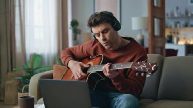 Gülümseyen gitarist evdeki kanepede dizüstü bilgisayarın kamerasında online ders kaydı yapıyor. Kulaklıkla gitar çalan yetenekli yakışıklı bir adam. Mutlu erkek müzisyen enstrümantal müzik yapıyor..