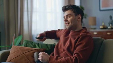 Gülümseyen adam TV 'de uzaktan kumandayla kanalı değiştiriyor. Mutlu adam en sevdiği televizyon dizisini bulduğu için seviniyor. Rahatlamış neşeli hipster dinlenme evinde kahve içiyor..