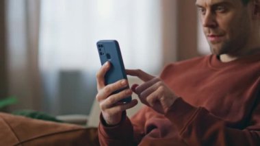 Yakın plan adam cep telefonunu araklıyor ve evdeki kanepede internetten alışveriş yapıyor. Rahatlamış milenyum adamı akıllı telefon ekranındaki koltuğa dokunuyor. Sakin ciddi erkek kaydırma haber kaynağı cep telefonu uygulamasında.