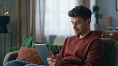 Evde tablet kullanarak internetten alışveriş yapan yakışıklı bir hippi. Rahat gülümseyen adam hafta sonu rahat koltukta bilgisayar ekranına dokunuyor. Apartmanda sosyal medyayı izleyen olumlu adam..