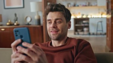 Daire kanepesinde cep telefonu ekranına bakan gülümseyen adam. Mutlu genç adam akıllı telefon mesajını evde okumaktan memnun. Yakışıklı serbest çalışan kapalı alanda cep telefonundan maaş bildirimi alıyor
