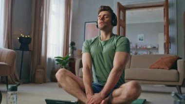 Huzurlu sporcu, evde yoga minderinde kablosuz kulaklıkla meditasyon yapıyor. Sakin adam gözlerini kapayıp tabletteki öğretici sesi dinliyor. Seren adam kulaklıkla nefes alma egzersizi yapıyor.
