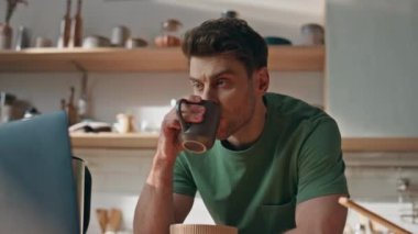 Sakin hipster mutfakta film izlerken kahve içiyor. Genç esmer adam dizüstü bilgisayar ekranında elinde sıcak çay fincanı tutuyor. Rahatlamış adam kahvaltıda evde vakit geçiriyor.