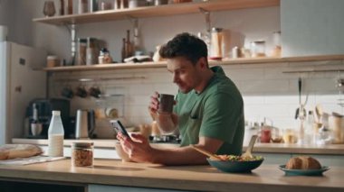 Ev mutfağında cep telefonuyla internette sörf yapan iş adamı. Akıllı telefondan mesaj okuyan mutlu, rahat adam dairede kahvaltıdan zevk alıyor. Gülümseyen adam internette gezinme uygulaması.