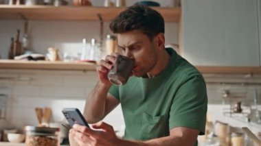 Odaklanmış bir adam mutfakta cep telefonu okuyarak kahvaltı yapıyor. Tıraşsız, ciddi bir adam elinde modern cep telefonuyla kahve içiyor. Sakin serbest çalışan öğle yemeğini tek başına yudumluyor.