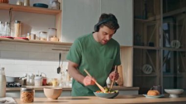 Kulaklıklı adam mutfakta en sevdiği şarkı boyunca yemek hazırlıyor. Komik neşeli adam, kablosuz kulaklık takıyor, müzik dinliyor sağlıklı sebzelerle salata pişiriyor. Yakışıklı erkek şarkı söylüyor.