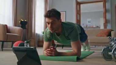 Sporcu, antrenman videosunu evde kapalı çekimde tablet eğitimiyle izliyor. Çevrimiçi dersi takip eden güçlü adam görünümlü bilgisayar ekranı. Formda sporcu dairede kalas egzersizi yapıyor.