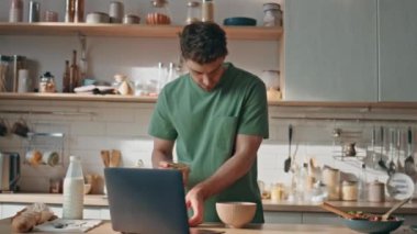 Yemek pişiren adam laptopla modern mutfakta kahvaltı hazırlıyor. Özel evde mesaj okuyan esmer bir adam. Evde bilgisayardan e-postaları kontrol eden konsantre yakışıklı bir erkek.