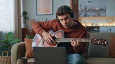 Dairedeki koltukta dizüstü bilgisayarla gitar çalmayı öğrenen odaklı bir gitarist. Evde gitar çalan, kablosuz kulaklıklı yetenekli bir adam. Yakışıklı adam kulaklıkla müzik dinleyip bilgisayara bakıyor..