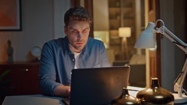 暗い家の閉鎖でPcでインターネットを閲覧する集中した男 現代のアパートでノートパソコンのキーボードの座席テーブルをタイプするハンサムな若者 コンピュータを使用してオンライン注文をする真剣な思慮深い顧客 — ストック動画