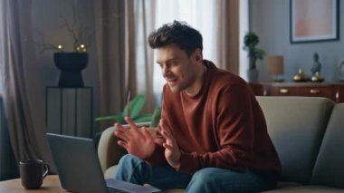 Konforlu dairedeki koltukta dizüstü bilgisayardan gülümseyen adam videosu. Rahatlamış yakışıklı adamın evde dostça bir toplantısı var. Oturma odasında bilgisayar kamerasına gülen genç bir erkek..