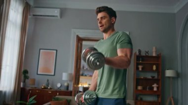 Apartman dairesinde dambıl egzersizi yapan gergin bir sporcu. Odaklanmış yakışıklı adam spor malzemelerini kaldırıyor güçlü eller kaslarını evde çalıştırıyor. Kas formundaki vücut için sağlıklı sporcu çalışması.