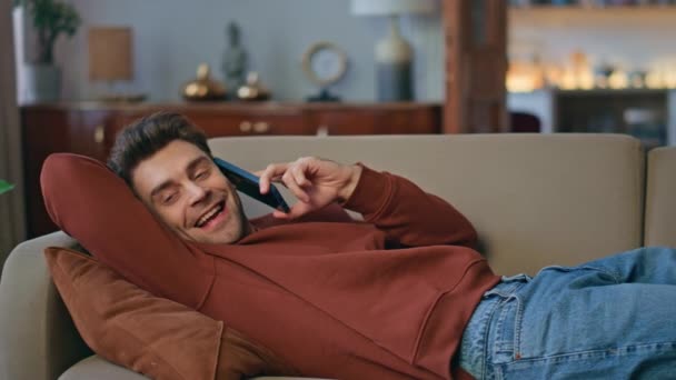 快乐的学生躺在舒适的沙发上聊天 积极英俊的男人喜欢在家里用智能手机进行友好的交流 年轻的自由撰稿人在国内周末与朋友交谈 — 图库视频影像