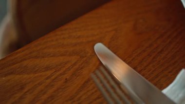 Çatal bıçağı modern kafeteryayı beyaz peçeteyle kapatmış. Zarif metal çatal bıçak seti ahşap masa örtüsü üzerinde konukların gelmesini bekliyor. Minimalist tasarımlı güzel bir kafe sofra takımı.