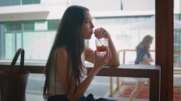 在近旁的现代街边餐馆里 一个放肆的女孩喝柠檬水 美丽的亚洲女人坐在木桌上 用稻草品尝美味的饮料 浪漫的女士在夏天的周末独自享受时光 — 图库视频影像