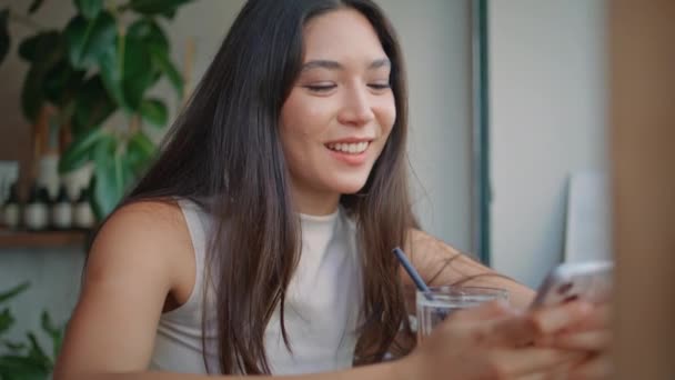 笑顔の学生少女が居心地の良いカフェで携帯電話をメッセージする ハッピータイの女性は レストランのテーブルに座っているスマートフォンでソーシャルメディアを閲覧します 携帯電話のスクリーンでSmsを読んでいる美しいアジアの女性 — ストック動画