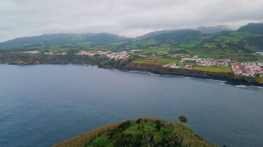 Bulutlu bir günde panoramik uçurum kıyısı kasabası. Mavi suyun kayalık yeşil pelerini yıkarken çekilen robot resmi. Kasvetli sabah havasında güzel, sakin bir sahil köyü. Huzurlu sahil dağları manzarası.