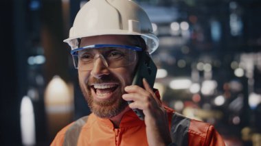 Endüstriyel üretim tesislerinde cep telefonuyla konuşan mutlu teknisyen. Gülümseyen sakallı mühendis akıllı telefondan harika haberler alıyor. Üniformalı profesyonel fabrika işçisi telefon ediyor..