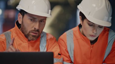 Dizüstü bilgisayar arayan mühendisler prodüksiyon projesini yakından kontrol ediyorlar. Koruyucu kask giyen iki fabrika işçisi fabrika tadilatını tartışıyorlar. Meşgul profesyonel teknisyenler