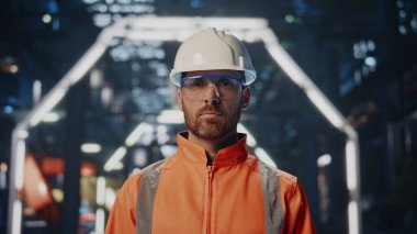 Yakından yetenekli fabrika işçisi üniformalı büyük üretim deposunda poz veriyor. Ciddi sakallı mühendis imalat tesisinde duruyor. Kasklı kendine güvenen profesyonel teknisyen kameraya bakıyor..