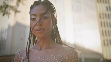 Afro örgülü kız rüzgarlı sokak portresi yapıyor. Şehvetli rastalı esmer, modaya uygun giyinmiş kameraya yakın çekim yapıyor. Kozmik stil sahibi kadın yaz melteminin tadını çıkarıyor güneş ışığı meydanında duruyor