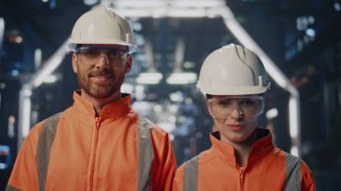 Modern yenilik üretiminde kameraya gülümseyen mutlu fabrika teknisyenleri. Üretim atölyesinde üniformalı pozlar veren yakın çift pozitif fabrika çalışanları. Koruyucu miğferler içinde duran mühendisler