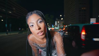 Dehşet saçan kız gece lambaları trafik ışıkları portresine bakıyor. Akşam bulvarında el kamerasıyla poz veren esmer tenli modacı kadın. Karanlık caddede modern kadın kollarını kavuşturuyor