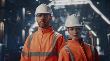 Modern endüstriyel makinelerin önünde poz veren kendinden emin iki imalat uzmanı. Gülümseyen bir çift mutlu fabrika mühendisi kameraya bakıyor. Fabrikada duran profesyonel teknisyenler..