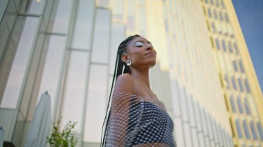 Neşeli esmer kıvrımlı cadde tek başına portre pozu veriyor. Güneşli şehir merkezinde şık giyinmiş, gülümseyen bir bayan kameraya bakıyor. Afro-Amerikan model yürüyen şehir kendini kaygısız hissediyor.