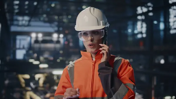 Engineer have work call at modern factory facility in protective hardhat uniform close up. Smart woman technician calling partners at production. Female workman discussing technical data by phone.