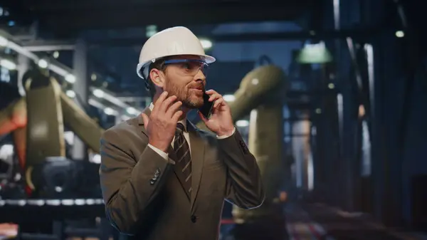 Smiling engineer talking smartphone at modern factory machinery in protective helmet closeup. Excited businessman speaking cellphone at heavy industry production. Manufacturing worker making work call
