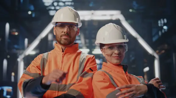 Professional team factory workers posing at modern heavy industry factory close up. Happy two technical specialists smiling standing manufacture workshop. Portrait proud engineers in protective helmet