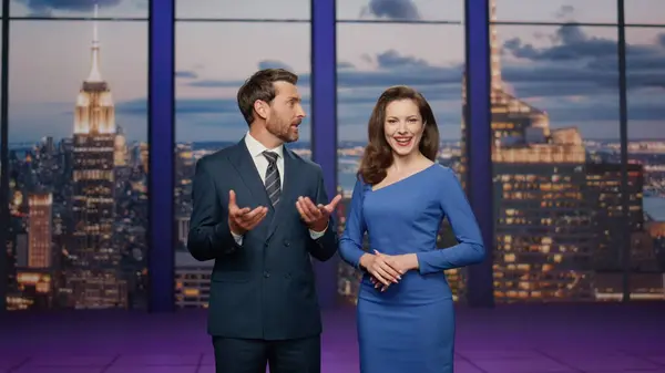 Friendly anchors smiling in studio television channel. Cheerful couple newsreaders reporting breaking news together lighting current information. Bearded anchorman talking to happy anchorwoman.