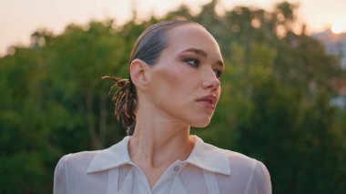 Yeşillik ağaçların önünde kendine güvenen bir modelin portresi. Yaz akşamında dışarıda poz veren güzel bir kadın. Kapatın. Beyaz tişörtlü güzel bayan hafta sonunun tadını çıkarıyor..