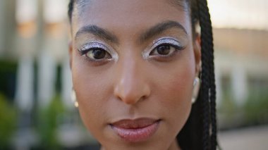 Şehir merkezindeki güneşli portrede muhteşem bir manken görünüyor. Makyajlı kozmik Afrikalı kadın yaz şehrinin yakın çekimlerinde poz veriyor. Dehşet saçan kadın tek başına yeşillik meydanına bakıyor.