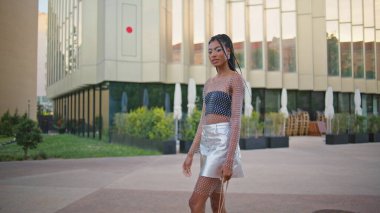Seksi esmer, tek başına yeşillik mahallesinde geziniyor. Şık giyinmiş, çantalı, boş sokaktan geçen mutlu Afro-Amerikan kadını. Moda mankeni kız yaz kasabasında sokakta yürüyor.