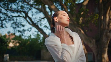 Yeşil Park 'ta yalnız başına güneşlenen bir kadın. Güneşli bahçede narin boynuna dokunarak poz veren çekici şehvetli bir model. Muhteşem moda kızı bahar doğasında huzuru hissediyor..