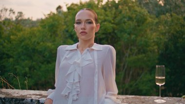 Ağaçların önünde duran kendine güvenen kadın yeşillik görünümlü kameraya yakından bakıyor. Dışarıda şarap kadehiyle poz veren çekici sakin modelin portresi. Rahat genç kız çitlere yaslanmış yeşil doğada rahatlıyor.