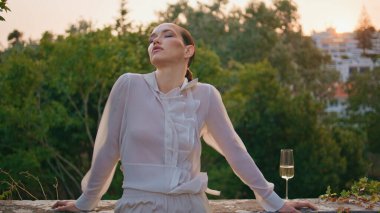 Şehvetli kız yaz akşamında rüzgarın esmesinden zevk alıyor. Havalı beyaz gömlekli baştan çıkarıcı kadın taş çitlere şarap kadehiyle yaslanıyor. Yeşillik ağaçların önünde poz veren zarif bir model..