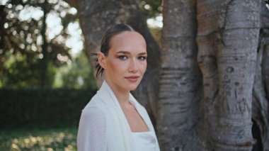 Muhteşem bayan yeşil yaz parkında yakından yürümeyi sever. Çekici zarif bir kadın bahçede geziniyor. Nazik bir gülümsemeyle kameraya bakıyor. Beyaz elbiseli romantik nazik kız ormanda sakinlik hissediyor..