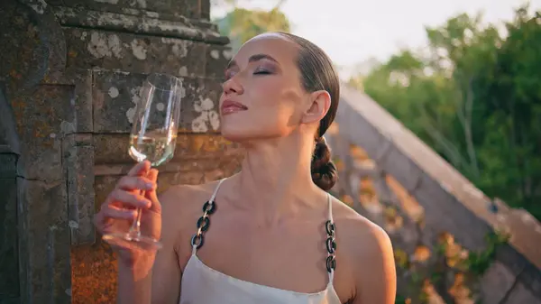 Relaxed girl drinking wine from glass goblet at old building staircase close up. Beautiful woman enjoy delicious alcohol closing eyes outdoors. Gorgeous lady looking camera standing in medieval castle